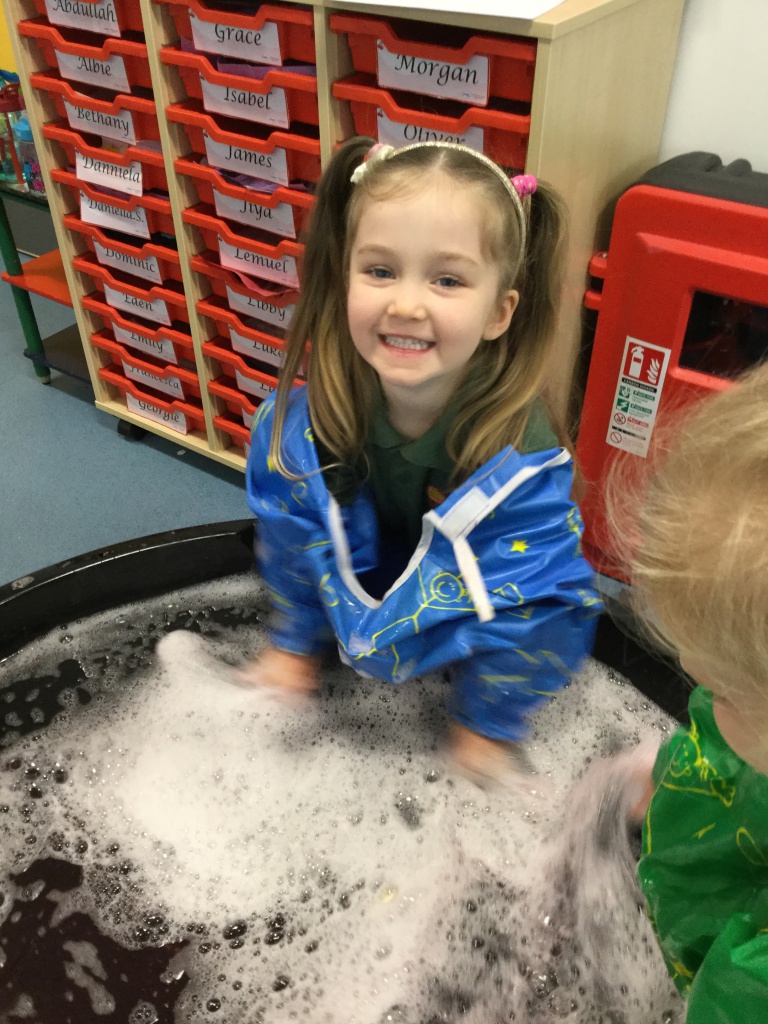 A young girl with pigtails smiling at the camera with her hands in soapy water.