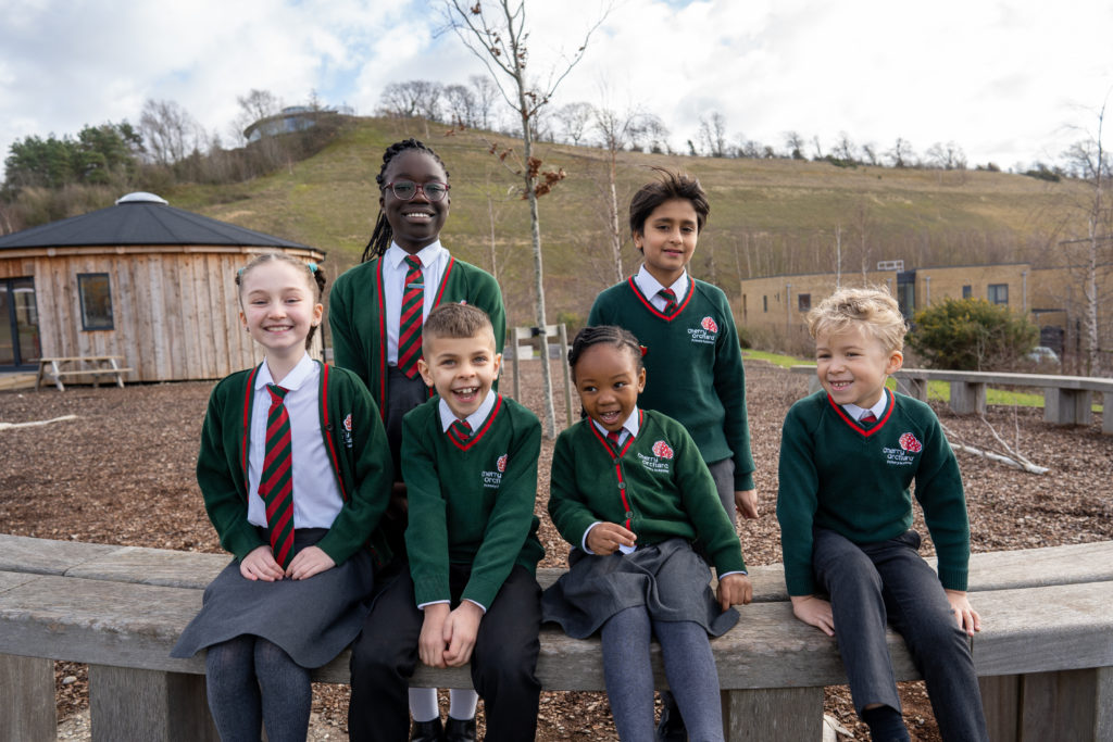 A group of students from Year 1 to Year 6 are seen smiling at the camera whilst sitting outdoors on a wooden bench.