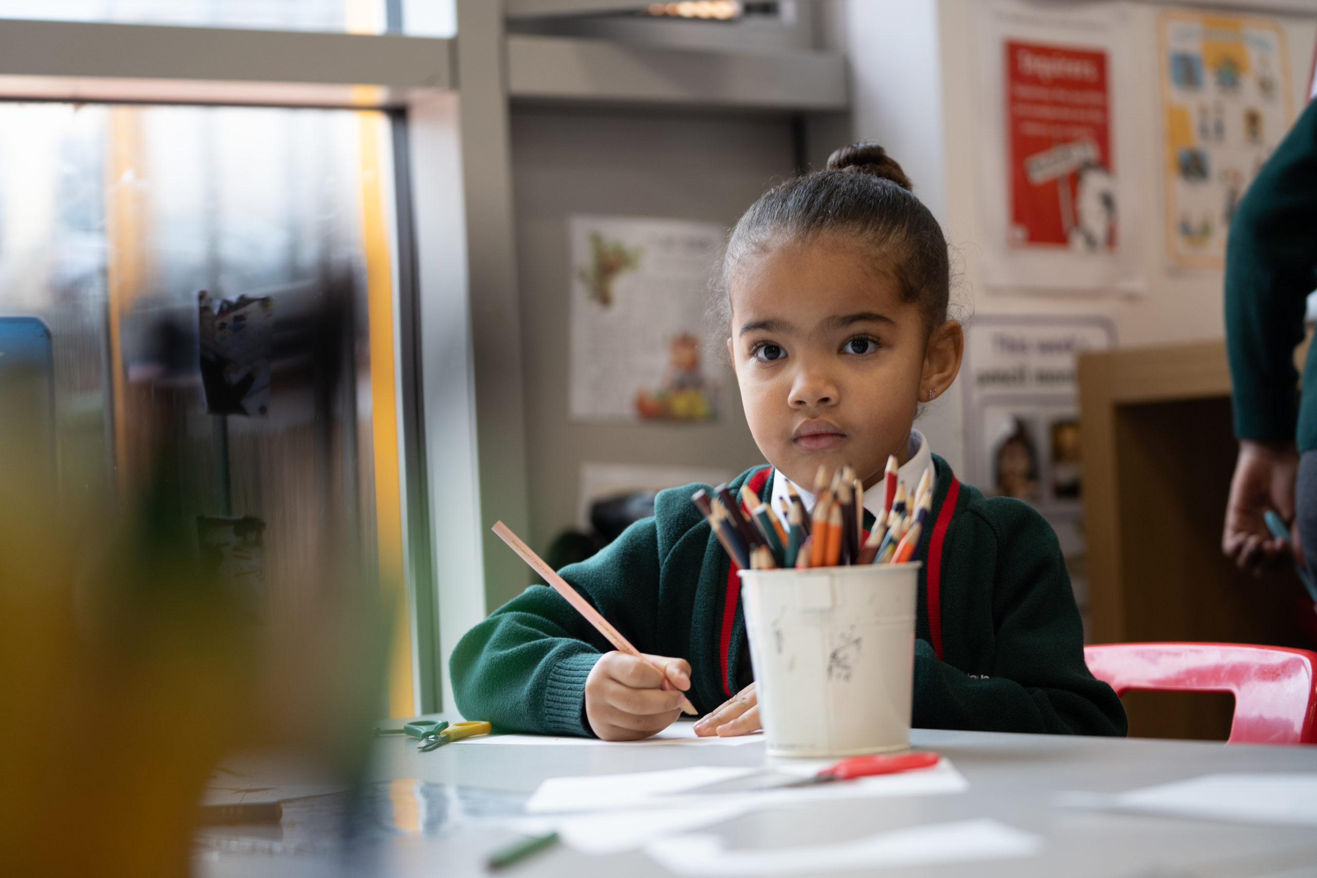 A young girl is shown sat at her desk, using coloured pencils to work on a piece of art she has created. She is looking directly at the camera.