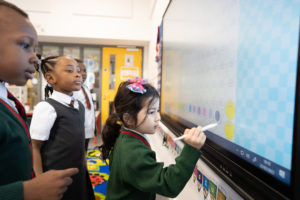 A small group of pupils are seen interacting with the Interactive Board at the front of the class to assist them with their learning.